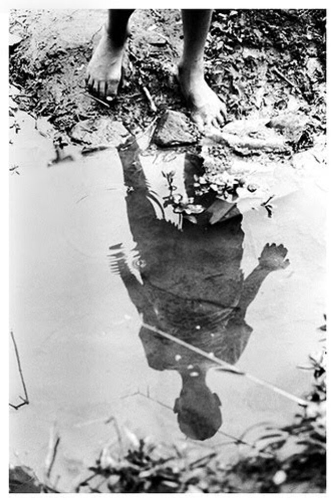 Fotografia em preto e branco de uma criança olhando o seu reflexo dentro de uma poção de água. 